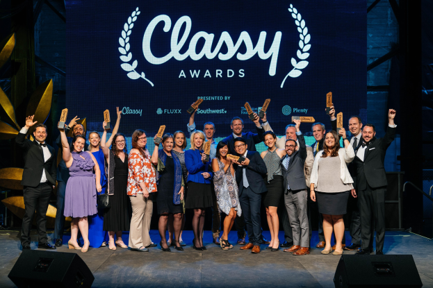 Eine Gruppe von Menschen auf einer Bühne mit Auszeichnungen, Lautsprechern darunter und einem Bildschirm mit der Aufschrift "Classy Awards 2019" im Hintergrund.