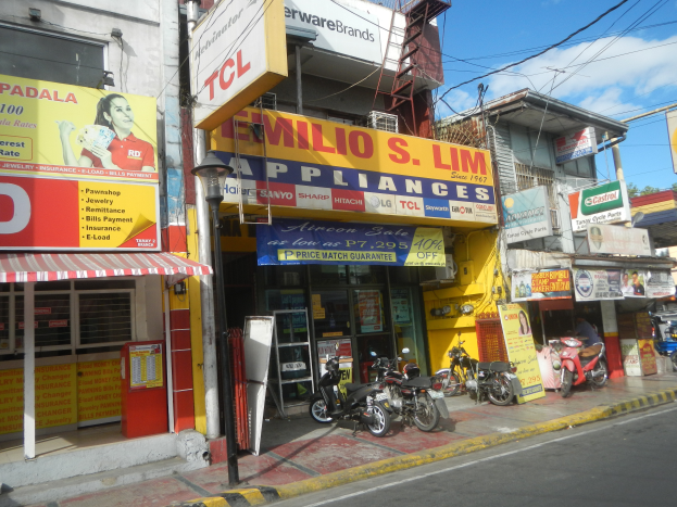 Eine belebte Straßenszene mit Gebäuden, geparkten Motorrädern, Straßenlaternen, Strommasten, Stromkabeln, Bäumen und einem bewölkten Himmel, mit einem Geschäft namens "Wilio S Lim Elektrogeräte" im Vordergrund.