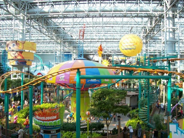 Großer Indoor-Wasserpark mit einer Achterbahn, umgeben von Menschen, Bäumen, Pflanzen und Deckenleuchten mit Stangen.
