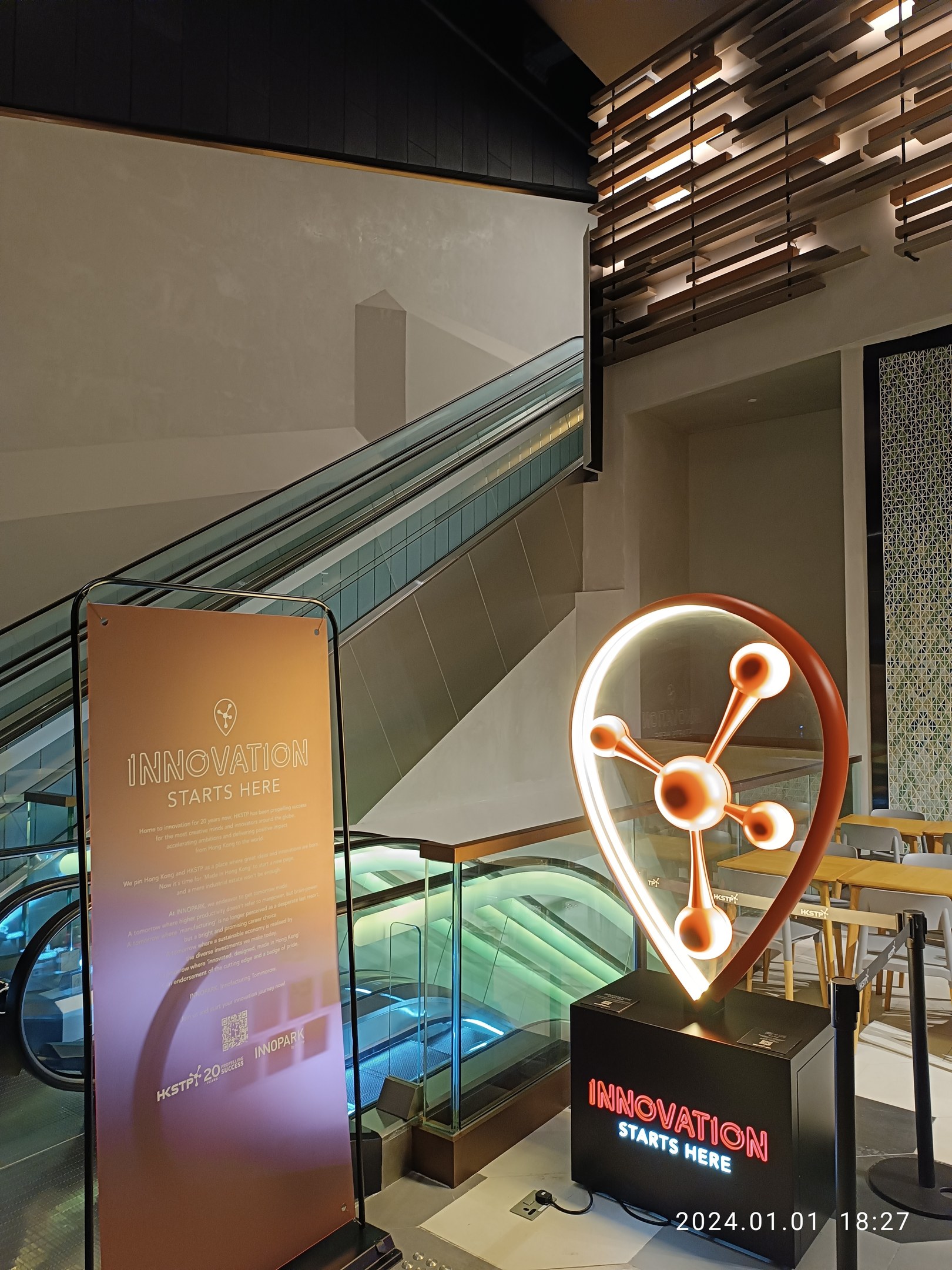 Großes Schild in einem Gebäude mit einer Rolltreppe im Hintergrund, flankiert von Tischen und Stühlen auf der rechten Seite und einer Tafel mit der Aufschrift "Innovation Starts Here" auf der linken Seite, was auf die HSTP20 Expo hinweist.