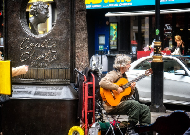 Ein Mann sitzt im Freien und spielt Gitarre, mit einer Statue links daneben, einem Baum im Hintergrund und einem weißen Auto und Geschäften rechts daneben.