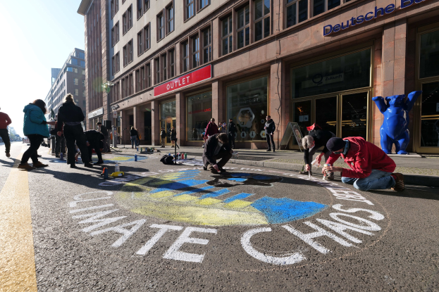 Eine Gruppe von Menschen sitzt vor einem Gebäude während einer Klimawandel-Demonstration in Berlin auf dem Boden, umgeben von Flaschen und anderen Gegenständen, mit Bäumen und einem klaren blauen Himmel im Hintergrund.