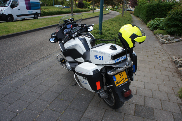 Ein polizeiliches Motorrad am Straßenrand mit einem Helm darauf, umgeben von Bäumen, Gebäuden und anderen Fahrzeugen.