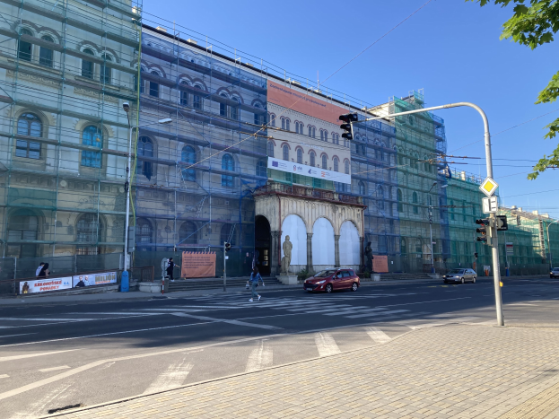 Großes Gebäude mit Gerüst während der Renovierung, umgeben von Fahrzeugen, Passanten, Ampeln, Schildern, Straßeninfrastruktur, Bäumen und einem klaren blauen Himmel.
