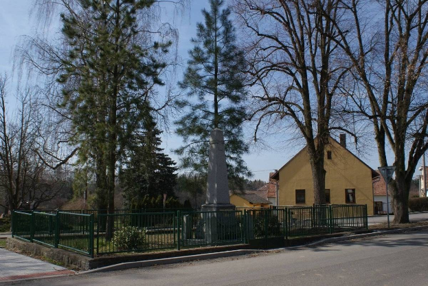 Ein Holocaust-Denkmal steht in der Mitte einer Straße, umgeben von einem Metallzaun, umgeben von Gras, Bäumen, Häusern, einem Straßenpfahl und einem bewölkten Himmel.