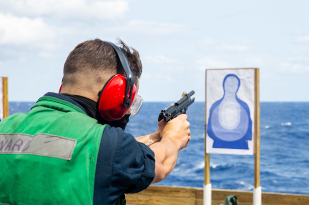 Ein Mann in einer grünen Jacke und roten Ohrschützern schießt mit einer Pistole auf ein Ziel, das vor einem hölzernen Geländer mit zwei Brettern steht, mit einem Meer und einem klaren blauen Himmel im Hintergrund.