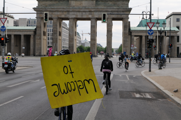 Eine Gruppe von Radfahrern mit Helmen fährt an der Brandenburgertor in Berlin vorbei, einer hält ein gelbes Schild, mit Laternenpfählen, Verkehrszeichen, Gebäuden, Bäumen und einem klaren Himmel im Hintergrund.