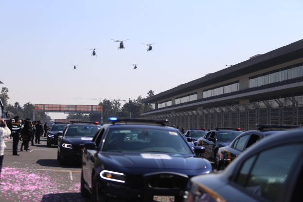 Polizeifahrzeuge vor einem Gebäude mit Menschen auf der linken Seite, Bäumen und einer Brücke im Hintergrund, Hubschrauber am Himmel unter einem klaren blauen Himmel.