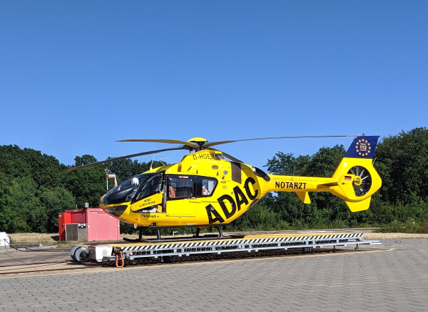 Ein gelber Hubschrauber steht auf dem Rollfeld eines Flughafens umgeben von Bäumen und einem klaren blauen Himmel, mit verschiedenen Gegenständen rund um den Hubschrauber.