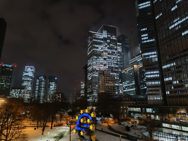 Europäischer Zentralbank-Hauptsitz in Frankfurt, Deutschland bei Nacht mit beleuchteten Gebäuden, Bäumen, Straßenlaternen, Lichtern und Fußgängern unter einem bewölkten Himmel mit Wasserzeichen.