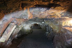 Ein langer, schwach beleuchteter Stollentunnel mit einem gewundenen Pfad, der zu einem Licht am Ende f√ľhrt, umgeben von hölzernen Stützen.