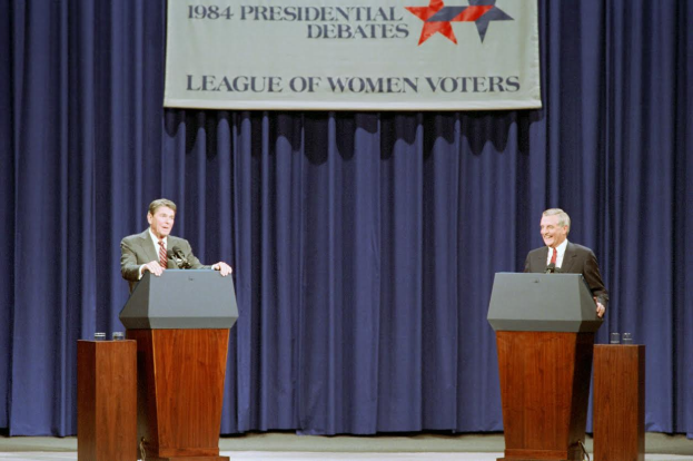 Zwei Männer in Blazern und Krawatten stehen an Podien mit Mikrofonen und Gläsern vor einem blauen Vorhang und einer Fahne mit Text und Sternen und diskutieren die 1984er Präsidentschaftsdebatten.