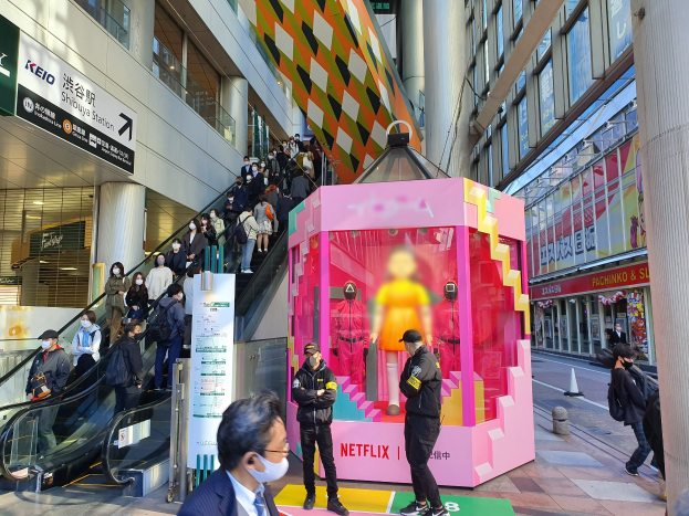 Eine Gruppe von Menschen steht vor einem Gebäude mit Glaswänden, Texttafeln, einige tragen Masken, in der Nähe einer Rolltreppe; ein pinker Kasten in der Mitte zeigt Schaufensterpuppen und trägt den Text "Netflix's new Netflix-Boot in Tokio."