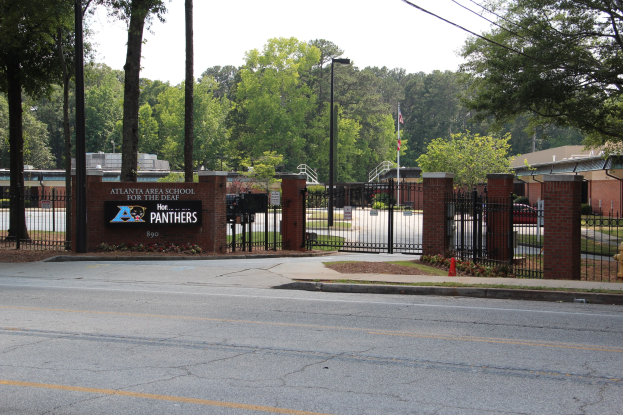 Eingang zu einer Schule mit einem Schild, umgeben von Bäumen, Straßenmöbeln, Fahrzeugen und Gebäuden unter einem klaren blauen Himmel.