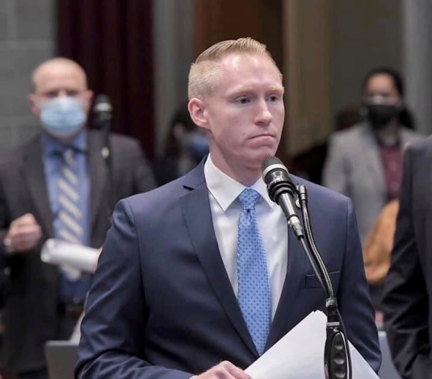 Ein Mann in Anzug und Krawatte spricht an einem Podium mit Papieren vor einem Mikrofon, während hinter ihm ein paar Menschen stehen und im Hintergrund eine Wand und ein Vorhang zu sehen sind.