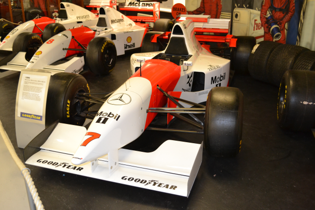 Eine Gruppe von Rennautos in einem Museum ausgestellt, umgeben von Menschen, ein Textschild, eine Seilabsperrung und eine Wand im Hintergrund.