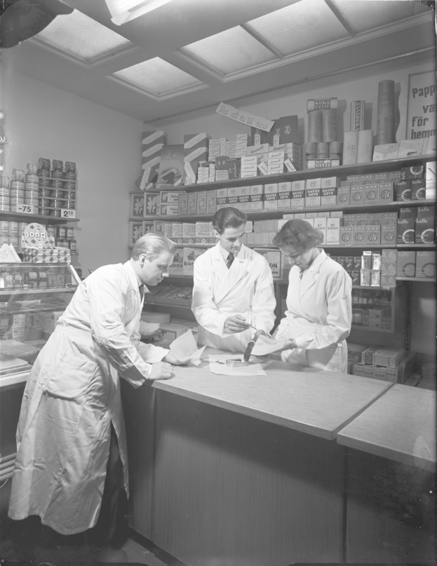 Drei Männer in weißen Kitteln an einer Apotheken-Theke mit Papieren und Gegenständen in einem Schwarz-Weiß-Bild.