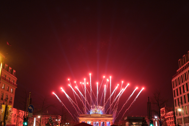 Eine nächtliche Stadtstraße in Berlin, Deutschland, mit dem Brandenburger Tor in der Mitte, umgeben von beleuchteten Gebäuden, Bäumen, Verkehrsampeln, Schildern und Flaggen, mit Feuerwerk am Himmel darüber.