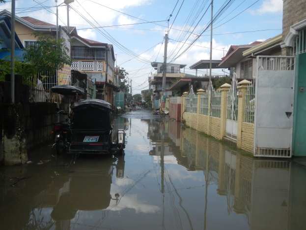 Eine überflutete Straße mit einem geparkten Motorrad, flankiert von Gebäuden, Bäumen, Polen und Drähten, unter einem bewölkten Himmel.