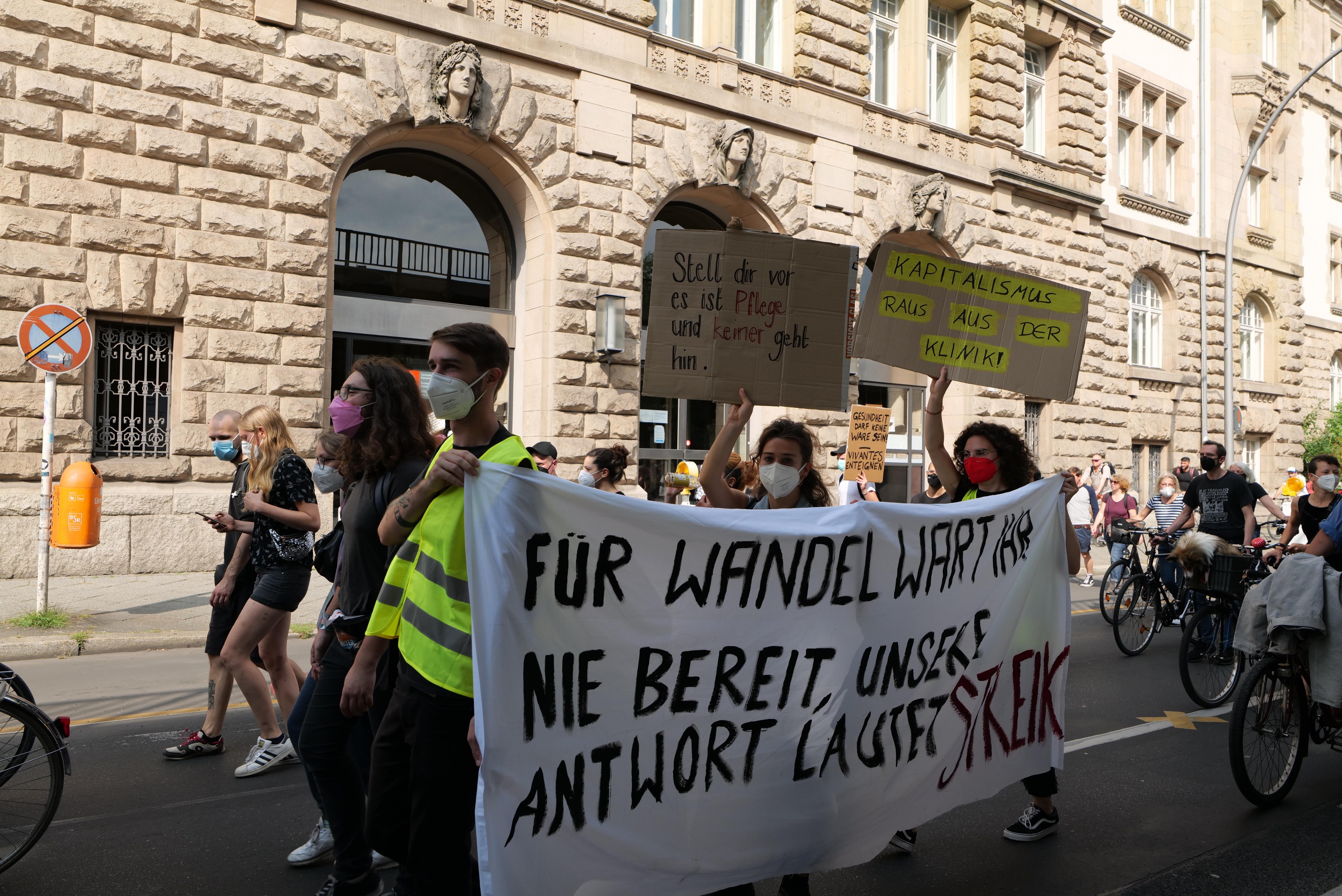 Eine Gruppe von Menschen, die auf einer Stadtstraße marschieren, einige halten Schilder und andere fahren Fahrräder, mit einem Gebäude mit Rundfenstern, Säulen und Skulpturen im Hintergrund.