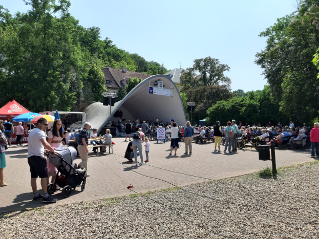 Eine Menschenmenge steht vor einem Gebäude bei der Festival-Eröffnung, einige sitzen auf Bänken, andere stehen; ein Kinderwagen ist im Vordergrund, Bäume und Gebäude sind im Hintergrund unter einem klaren blauen Himmel, Zelte sind links und ein Mülleimer rechts.