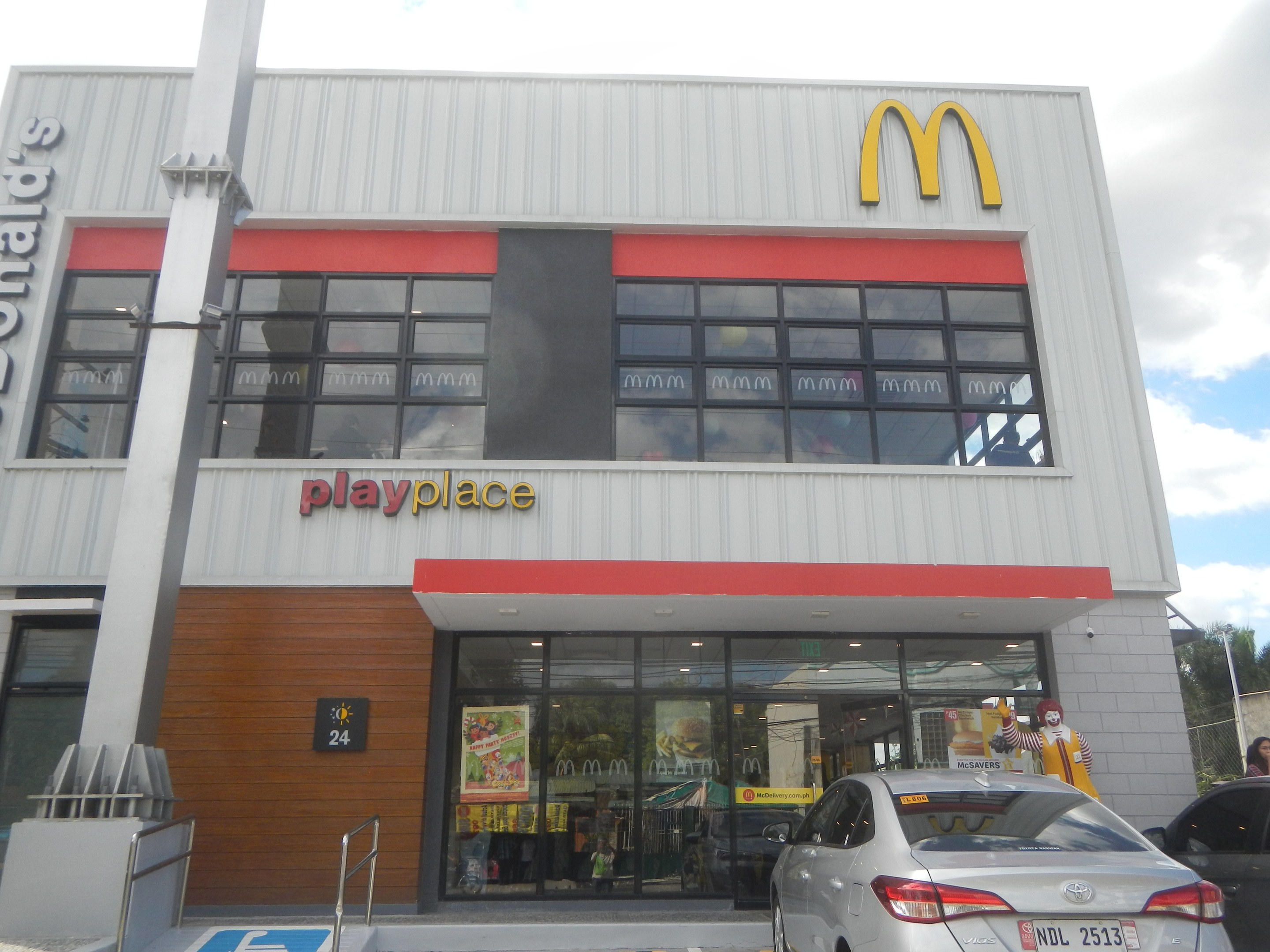 Ein McDonald's-Restaurant mit parkenden Autos, umgeben von Bäumen und Strommasten unter einem klaren blauen Himmel, mit Glaswänden, einem "McDonald's PlayPlace"-Schild und sichtbaren Innenpostern und Gegenständen.