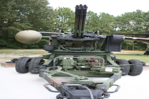 Militärfahrzeug mit montierter großer Kanone in einem Freilichtmuseum ausgestellt, unter einem klaren blauen Himmel und umgeben von Bäumen.