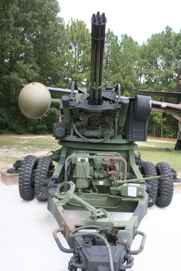 Militärfahrzeug mit montierter großer Kanone in einem Freilichtmuseum ausgestellt, unter einem klaren blauen Himmel und umgeben von Bäumen.
