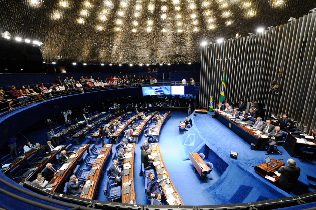 Eine Konferenzbühne mit sitzenden Personen an einem Tisch mit Papieren und Gegenständen, flankiert von Fahnen und einer Skulptur im Hintergrund, mit Zuschauern an Tischen und hinter einer Absperrung mit Bildschirmen, unter dekorativen Deckenleuchten.