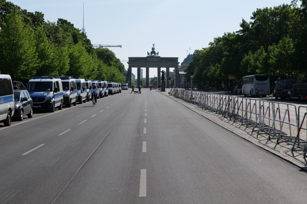 Lange Reihe von Polizeiwagen auf einer Straße vor dem Brandenburger Tor geparkt, mit Fahrradfahrern und Füttern, Barrieren, Bäumen und einem Bogen mit Statuen im Hintergrund.