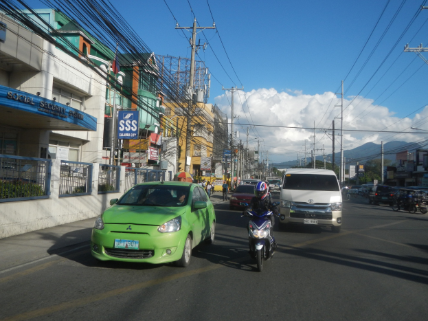 Ein grünes Auto fährt eine Straße entlang, flankiert von hohen Gebäuden mit Strommasten und -leitungen darüber, Fußgängern auf dem Gehweg und Bäumen, Bergen und Wolken im Hintergrund.