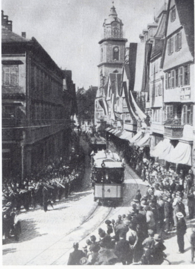 Schwarzes und weißes Foto einer Stadtstraße mit einer Tram in der Mitte, flankiert von Gebäuden mit Fenstern, und einem Kirchturm im Hintergrund; Text unten.
