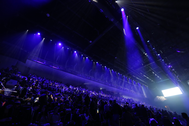 Eine große Menschenmenge sitzt in einer Konzertarena und schaut auf eine Bühne mit Deckenlampen und einem Bildschirm auf der rechten Seite.