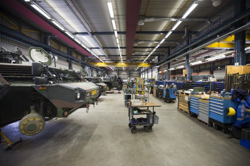 Großes Lager mit Maschinen, Fahrzeugen links, Wagen mit Gegenständen und Holzboxen rechts sowie Deckenbeleuchtung.