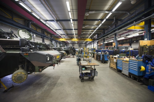 Großes Lager mit Maschinen, Fahrzeugen links, Wagen mit Gegenständen und Holzboxen rechts sowie Deckenbeleuchtung.