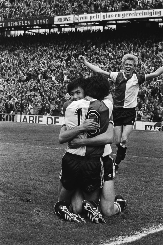 Drei Fußballspieler umarmen sich auf dem Boden und feiern ein Tor vor einem jubelnden Publikum in einer schwarz-weißen Stadionszene.