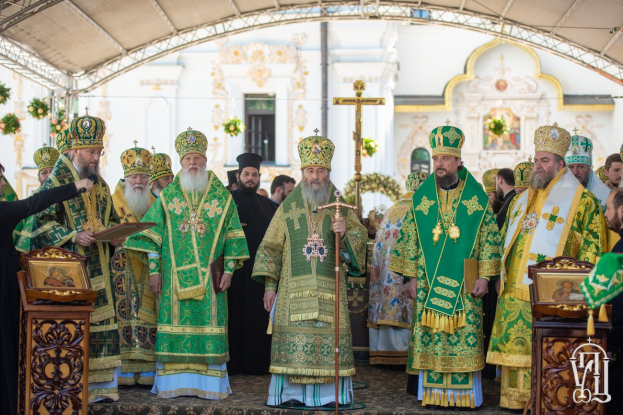 Gruppe von Priestern vor einer Kirche während einer religiösen Zeremonie, einer Person mit einem Buch und einem Mikrofon auf der linken Seite des Bildes, einem Kreuzsymbol und Blumen im Hintergrund.