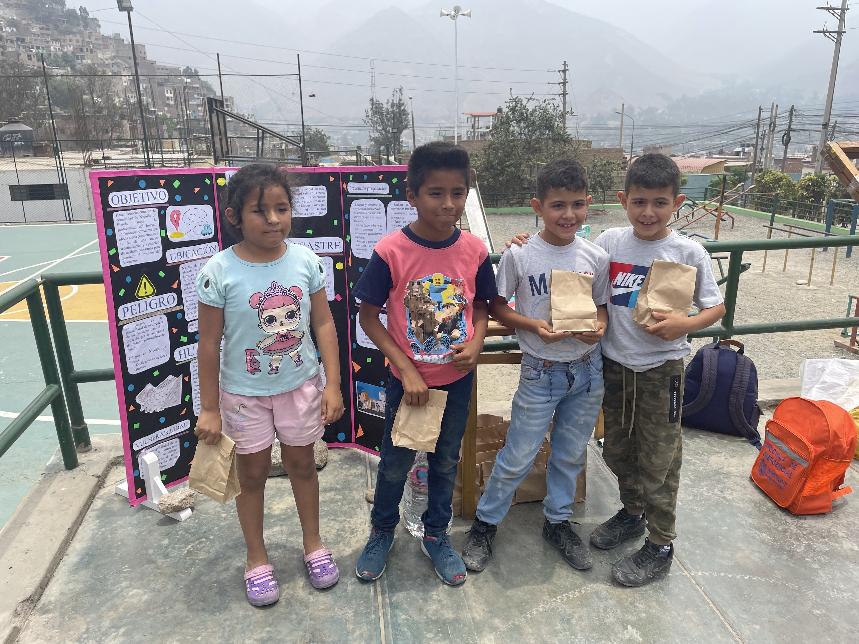 Gruppe von Kindern auf einem Basketballfeld mit Papiertüten in den Händen, mit Vorräten auf dem Boden und einer Tafel mit Text links daneben, vor dem Hintergrund von Zäunen, Pfosten, Straßenlaternen, Bäumen, Gebäuden, Hügeln und Himmel.