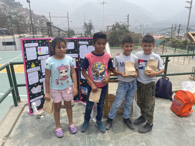Gruppe von Kindern auf einem Basketballfeld mit Papiertüten in den Händen, mit Vorräten auf dem Boden und einer Tafel mit Text links daneben, vor dem Hintergrund von Zäunen, Pfosten, Straßenlaternen, Bäumen, Gebäuden, Hügeln und Himmel.
