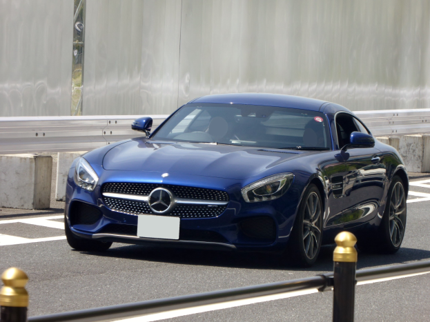 Blauer Mercedes-Benz AMG GT Coupé mit schlanker, aerodynamischer Gestaltung, das eine Straße entlangfährt, mit eckigen Scheinwerfern, einem großen Kühlergrill, glänzenden großen Rädern und einem luxuriösen Lederinnenraum mit einem modernen Armaturenbrett, vor einer Wand im Hintergrund.