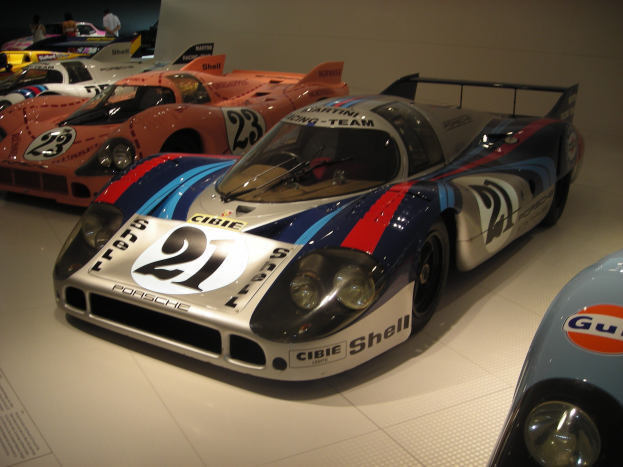 Ein Porsche 917K Rennwagen aus Le Mans in einem Museum ausgestellt, umgeben von anderen Sportwagen, mit Menschen in der Nähe und einer Wand im Hintergrund; ein Papier mit Text ist links sichtbar.