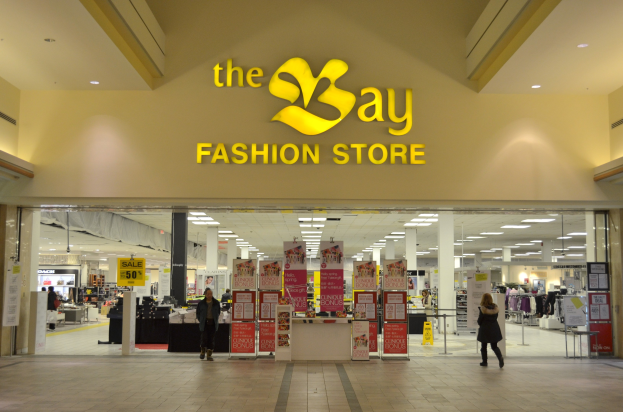 Außenansicht eines Bay Fashion Store in einem Einkaufszentrum mit Passanten, Wandtextanzeigen, Säulen, Deckenleuchten und Hintergrunddekoration.