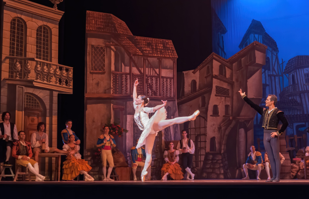 Zwei Tänzer performen auf einer Bühne im Royal Opera House in London, mit einem Publikum im Vordergrund und einer Leinwand mit Gebäudeabbildungen im Hintergrund.