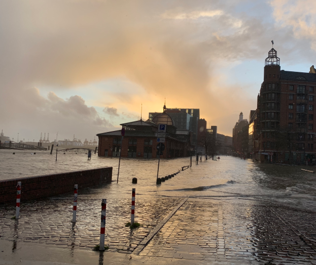 ├ťberflutete Stra├če in Hamburg, Deutschland, mit Wasser auf der Stra├če, Masten, Schildern, Geb├Ąuden mit Fenstern, einer Br├╝cke und einem bew├Âlktem Himmel im Hintergrund.
