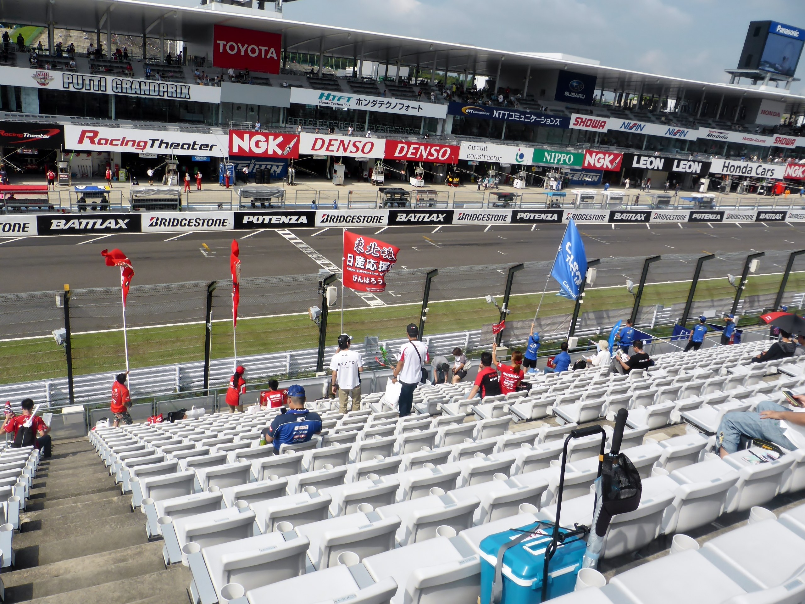 Eine Gruppe von Menschen sitzt auf Tribünen in einem Stadion, mit Fahnen, Texttafeln, Geländern und Stufen, unter einem bewölkten Himmel.