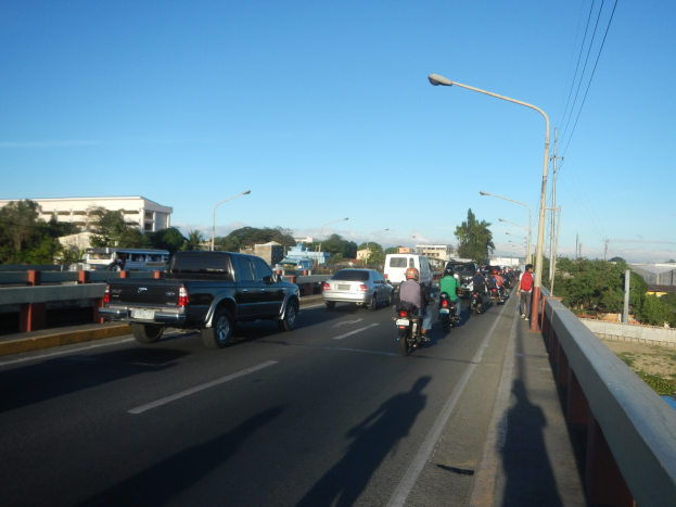 Eine Gruppe von Motorradfahrern überquert eine Brücke mit einem Pick-up-Truck im Vordergrund, Bäume und Gebäude sind im Hintergrund unter einem klaren blauen Himmel zu sehen.