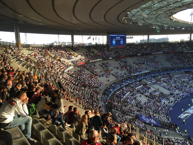 Große Zuschauermenge in einem Stadion bei einem Fußballspiel mit einer Bühne, Fahnen, Stangen, einem Bildschirm und der Allianz Arena in München, Deutschland im Hintergrund.