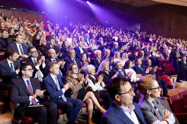 Eine große Gruppe von Menschen in einem Auditorium, die klatschen, wobei einige Kameras und Scheinwerfer zu sehen sind.