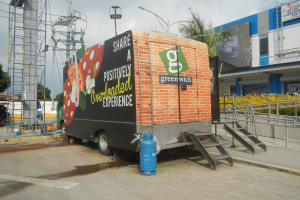 Lkw vor einem Gebäude mit einer Tafel, auf der "Teilen Sie eine positiv überladene Erfahrung" steht, mit Stufen, einem Zylinder auf dem Boden, Geländern, Strommasten mit Drähten, Laternen, Bäumen und einem bewölkten Himmel.