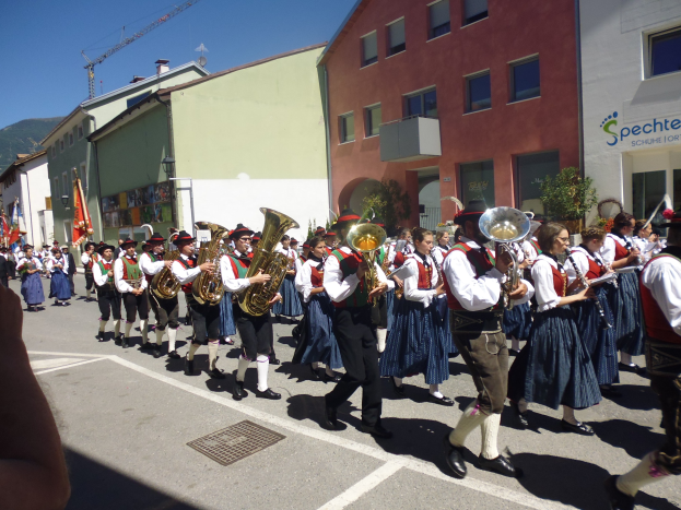 Gruppe von Menschen in traditioneller bayrischer Tracht, die auf einer Straße mit Gebäuden Musikinstrumente spielen, einige halten Fahnen, mit einem Hügel und einem klaren blauen Himmel im Hintergrund.